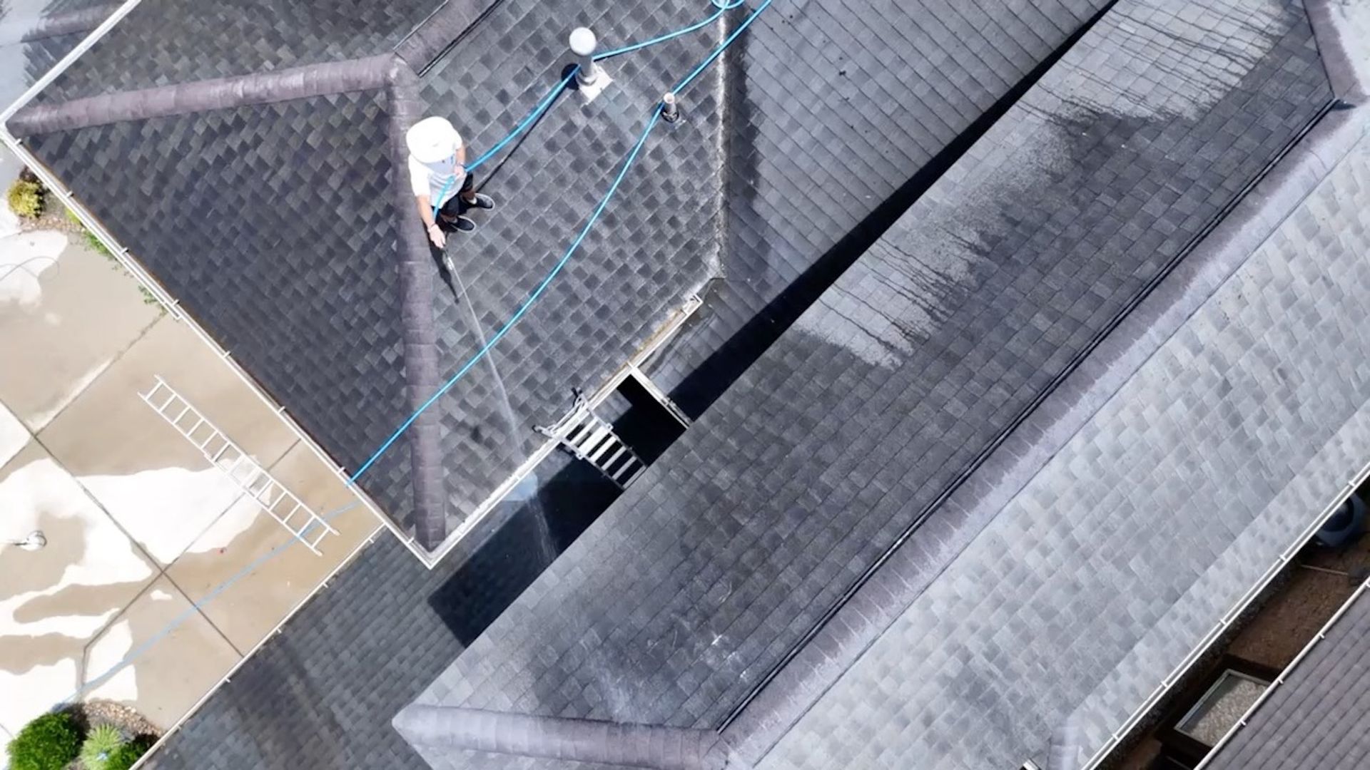 Roofers working on a gray shingled roof beside a steep roof valley and chimney.