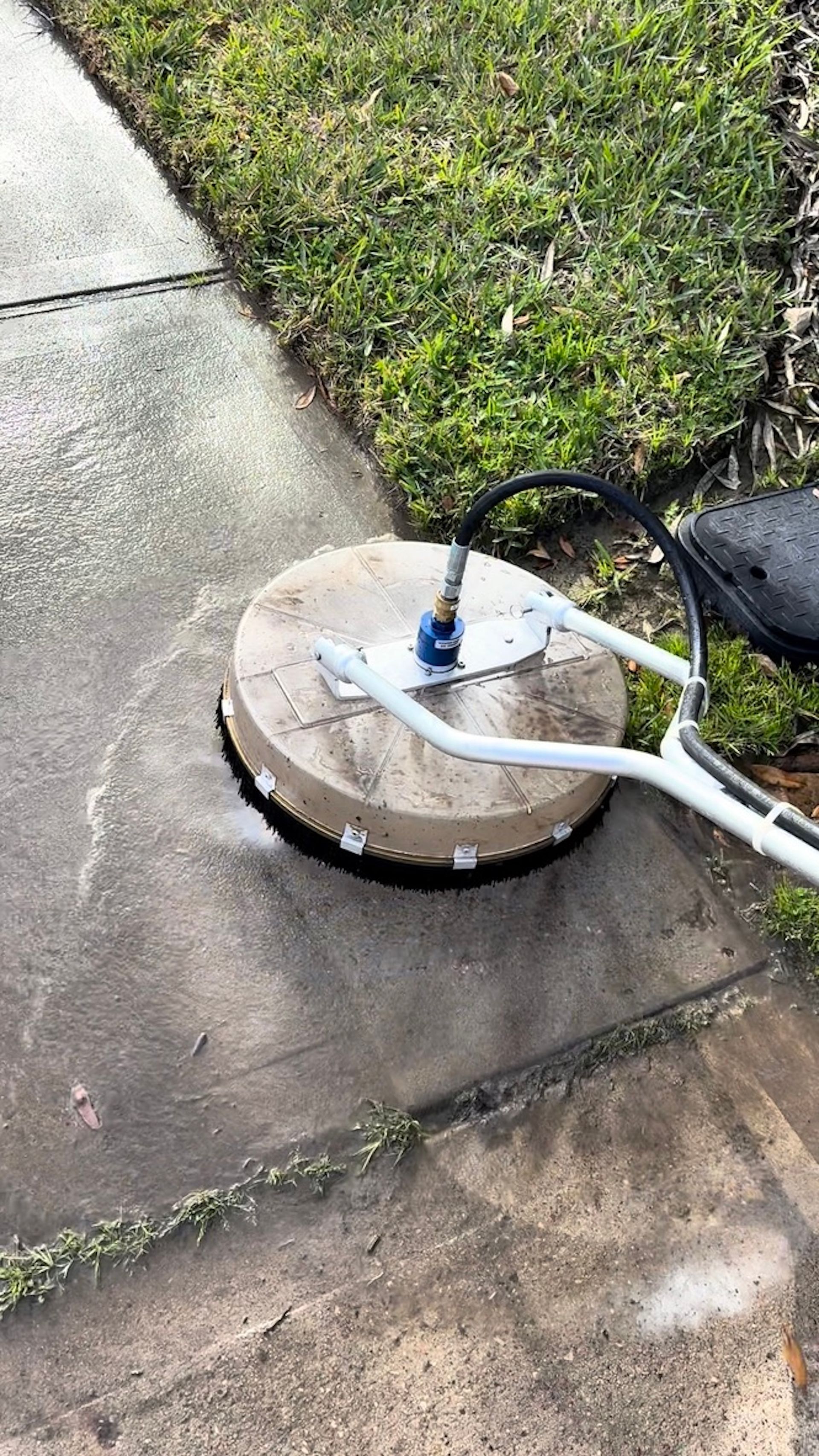 Round concrete utility cover beside a sidewalk, with white hoses attached on a grassy edge.