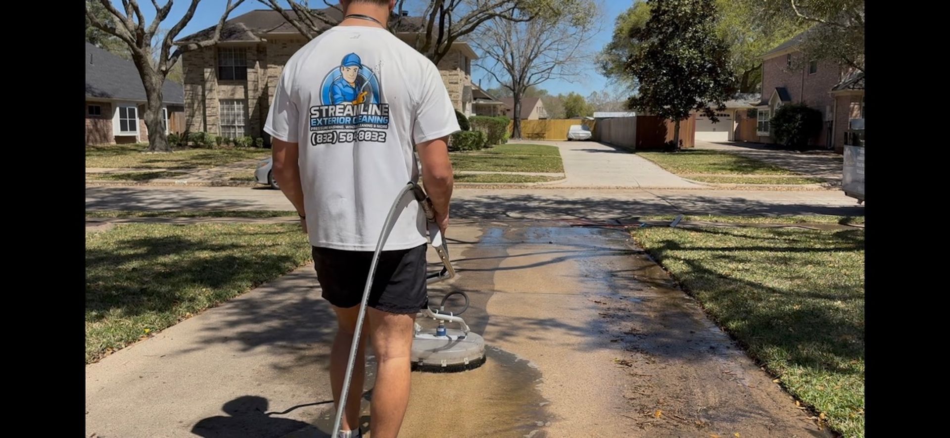 Person power washing a sidewalk in a suburban neighborhood, spraying water along a wet concrete path.