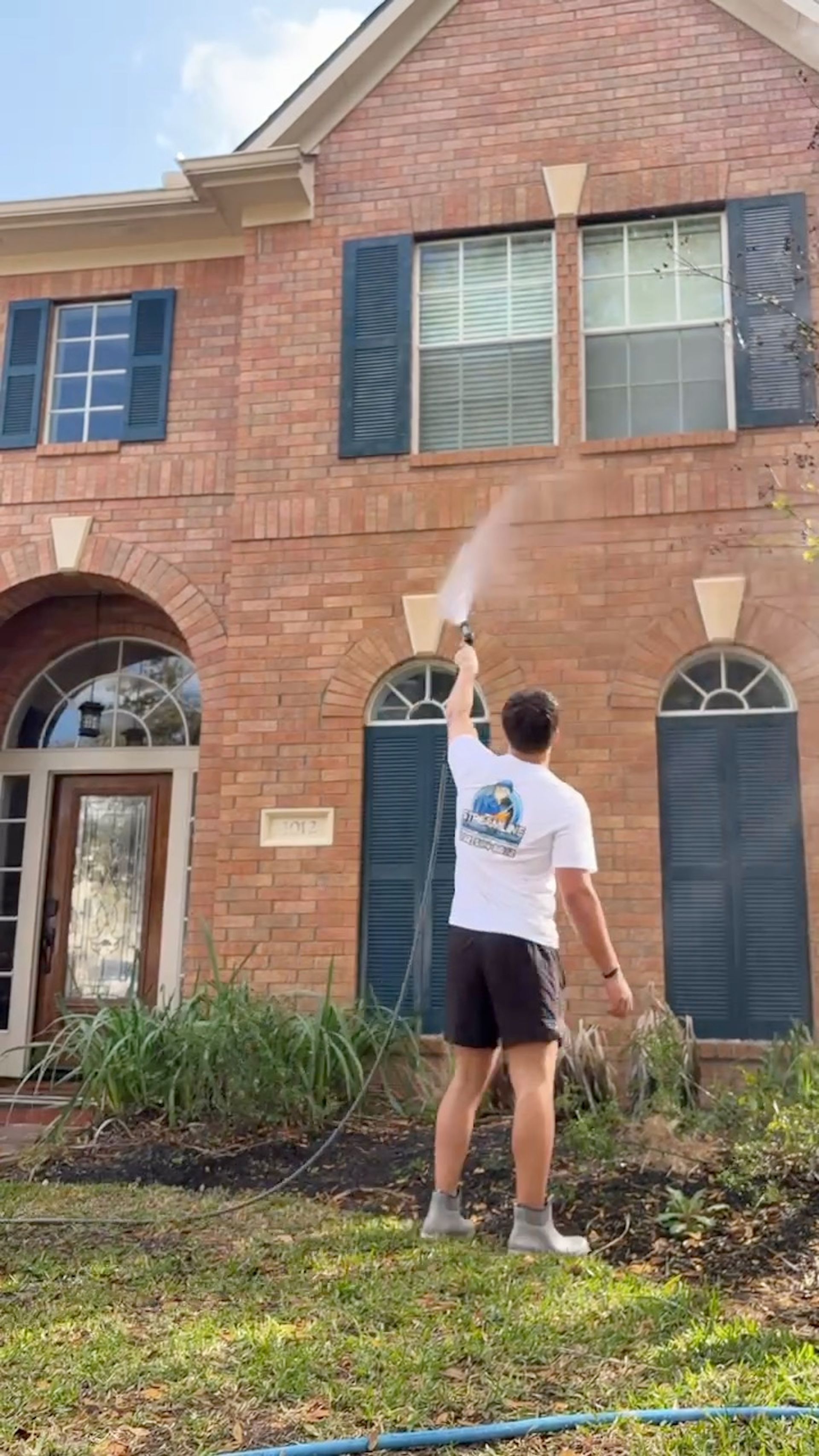Person painting a two-story brick house exterior with a roller on an extension pole.