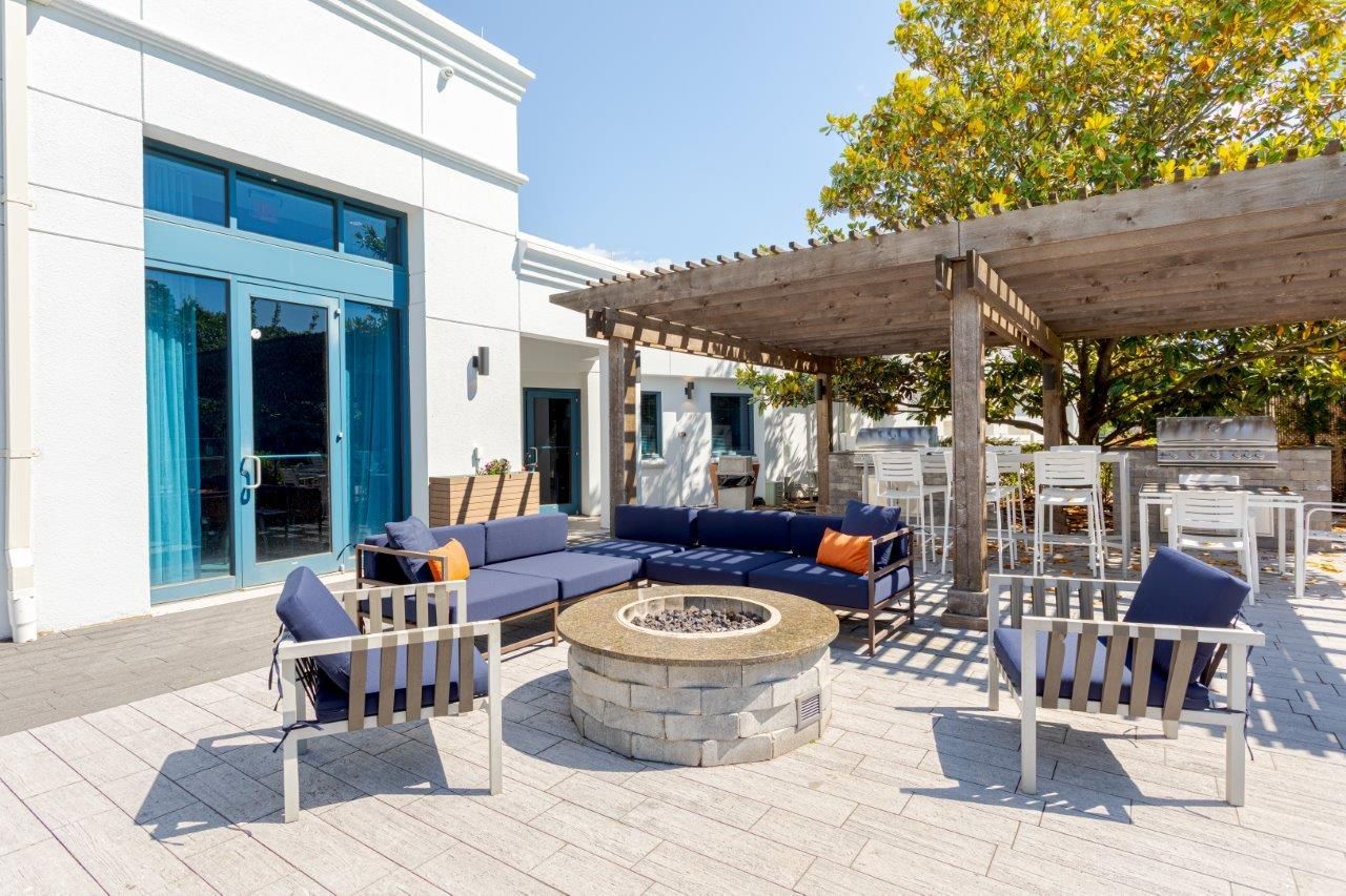 Outdoor patio with blue sofas, white chairs, a fire pit, and a pergola beside a white building