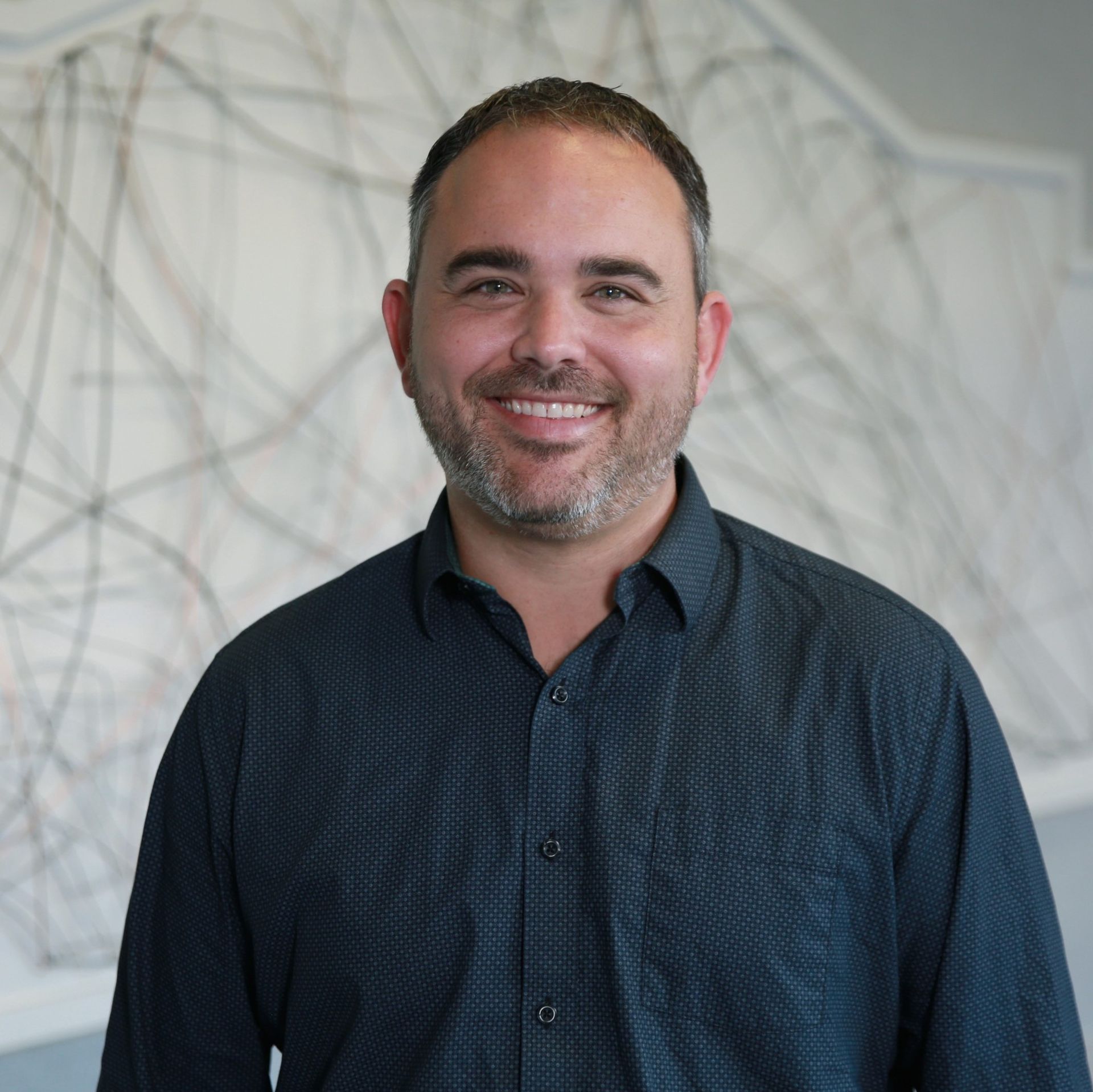 Man with a beard smiling, wearing a dark button-down shirt, standing in front of an abstract art piece.