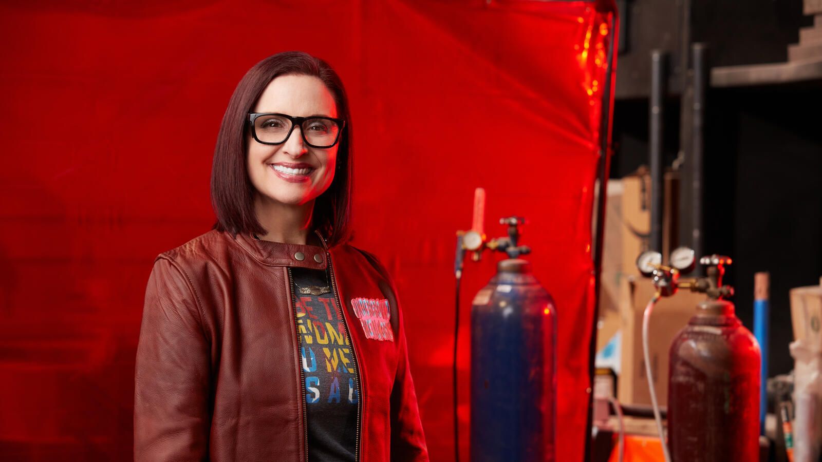Woman with glasses smiles in a red-lit workshop, near welding equipment. She wears a leather jacket and graphic tee.