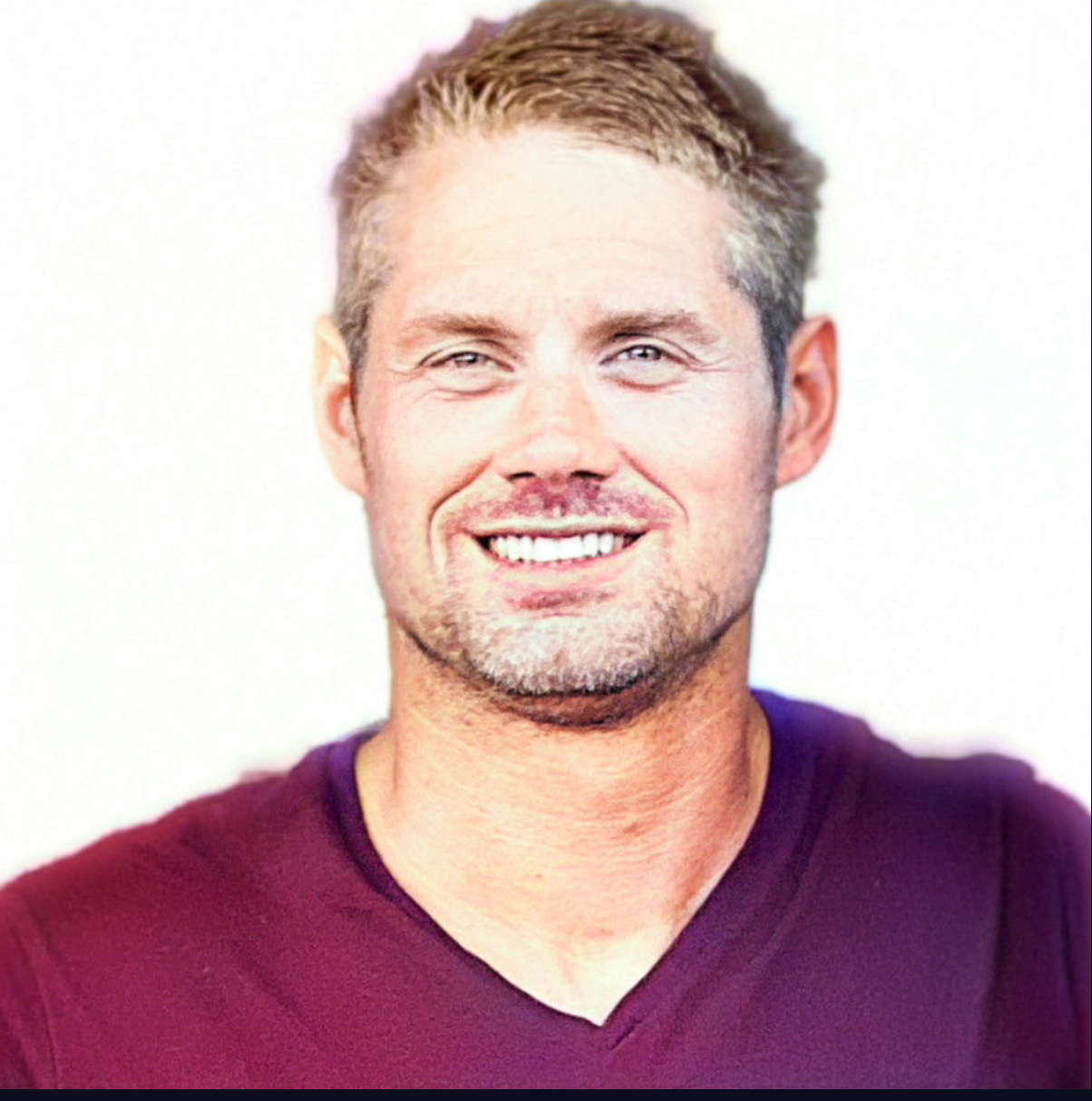 Man with blond hair smiling, wearing a burgundy v-neck shirt, against a white background.