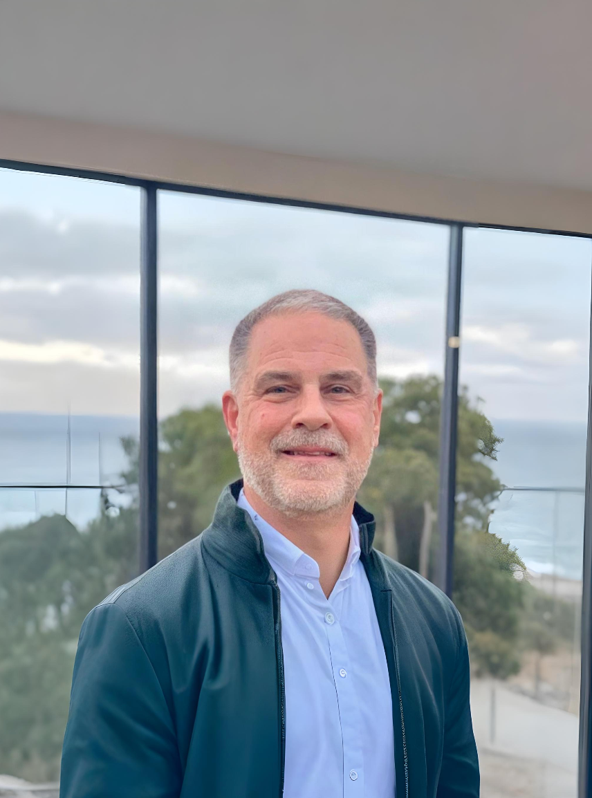 Man in teal jacket and light blue shirt smiles near large windows with a scenic view.