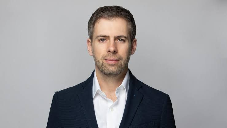 Man in a white shirt and navy blazer looks at the camera against a gray background.