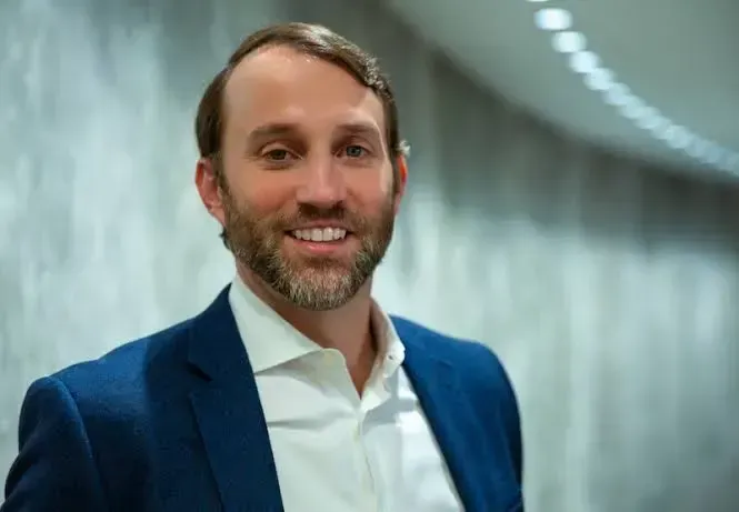 Man in blue blazer smiles, standing in a hallway.