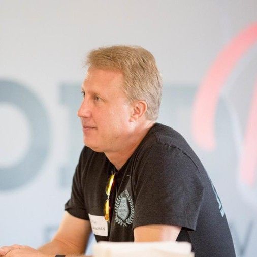 Man in black t-shirt, wearing a name tag, looking to the side, seated.