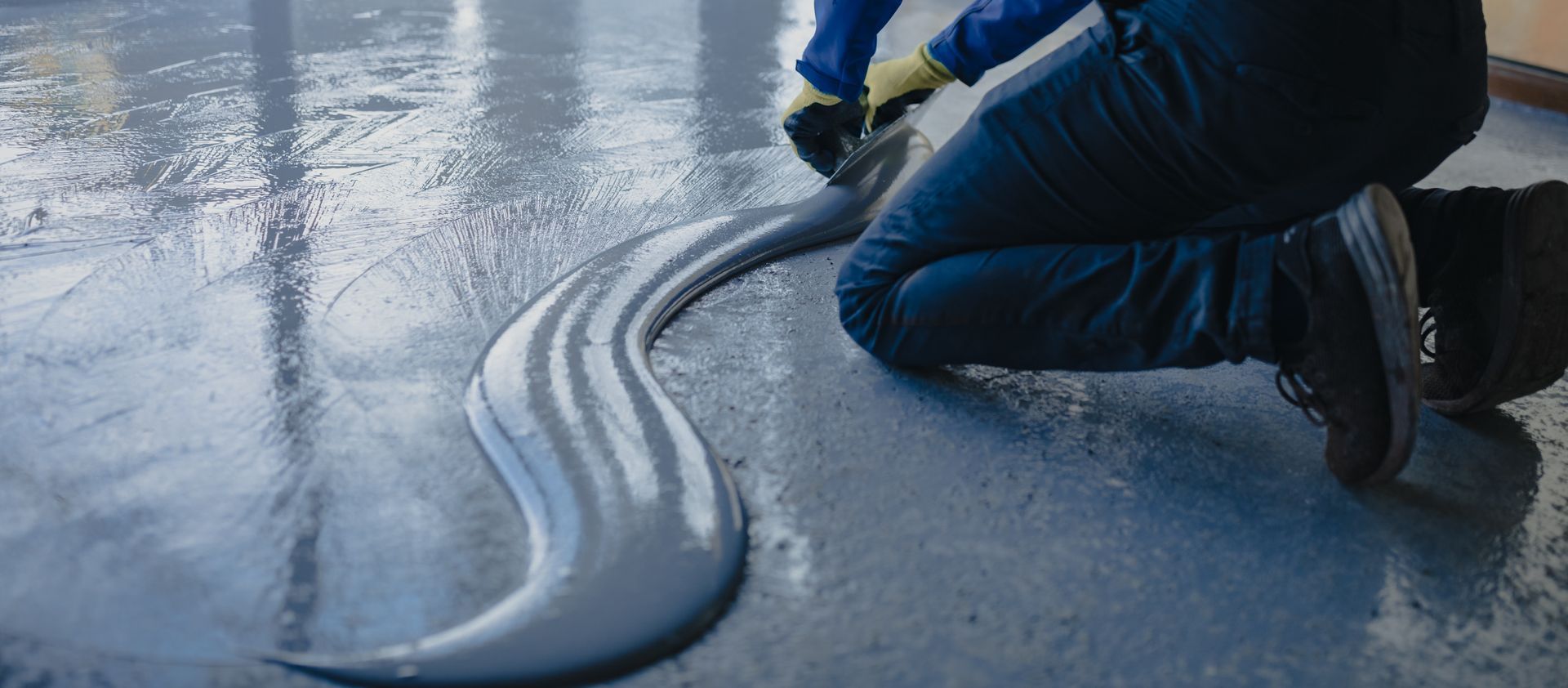 Worker kneeling on icy pavement, spreading a thick gray sealant or coating from a hose
