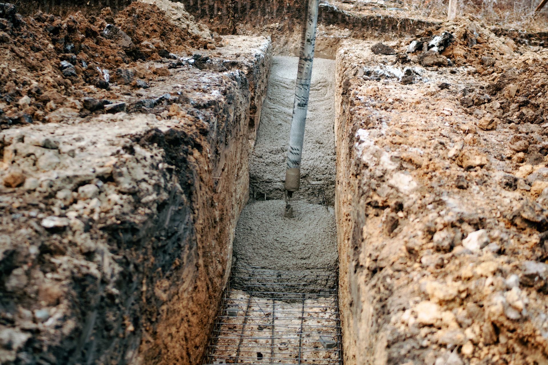 Narrow trench with concrete footing being poured, flanked by rough dirt walls and muddy ground