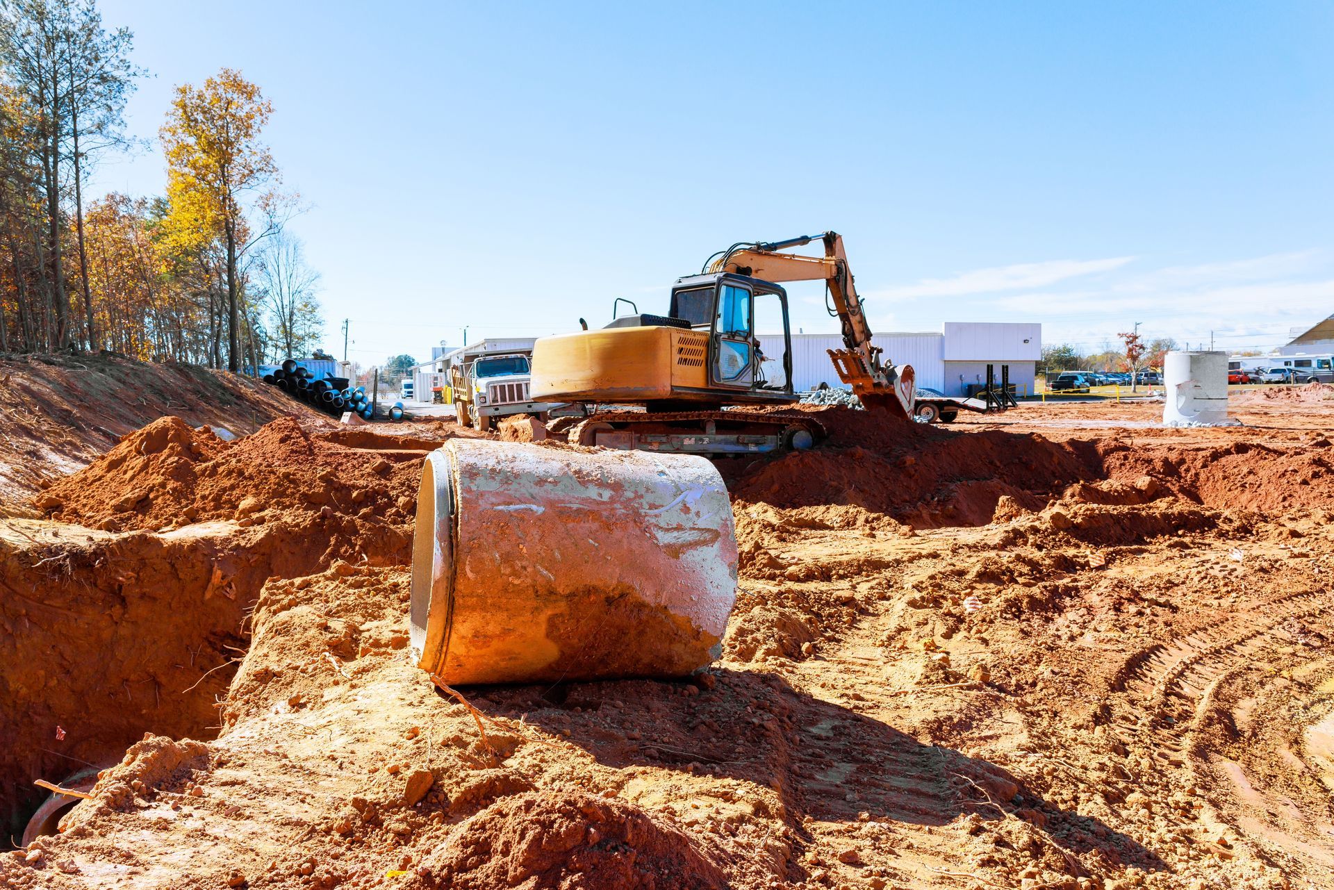 Excavator at a red dirt construction site with a large metal pipe in the foreground under a clear blue sky