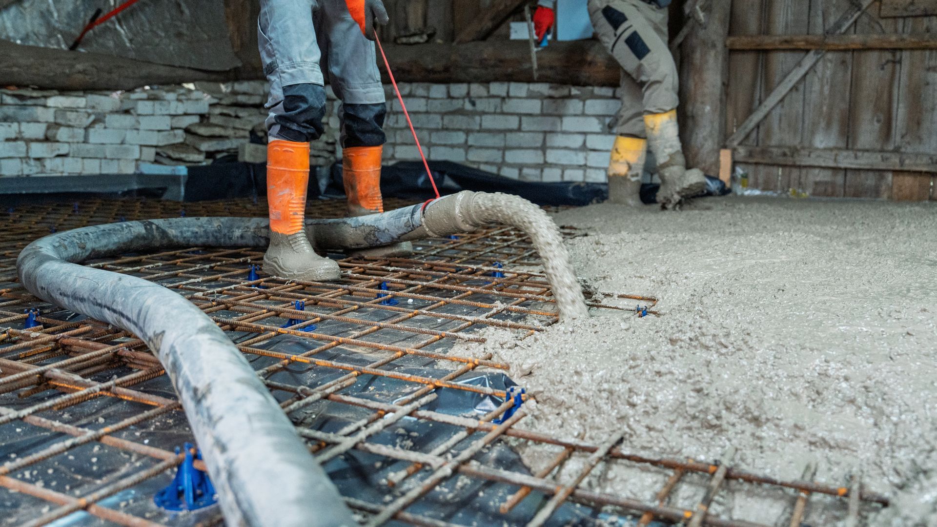 Workers in orange boots pour concrete over rebar at a construction site