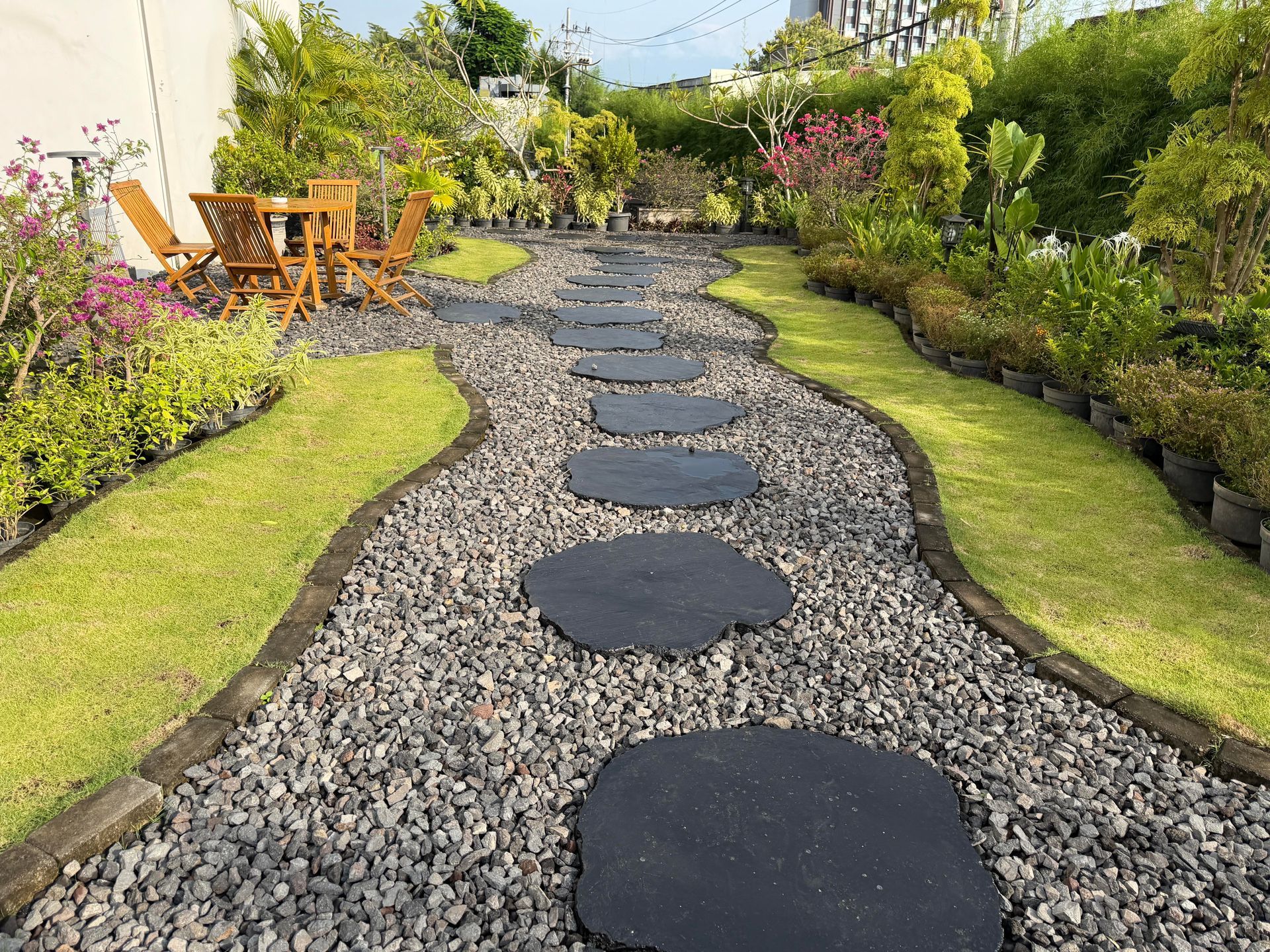 Curved stepping-stone path through a lush garden with green lawns, flowers, and wooden chairs in the background