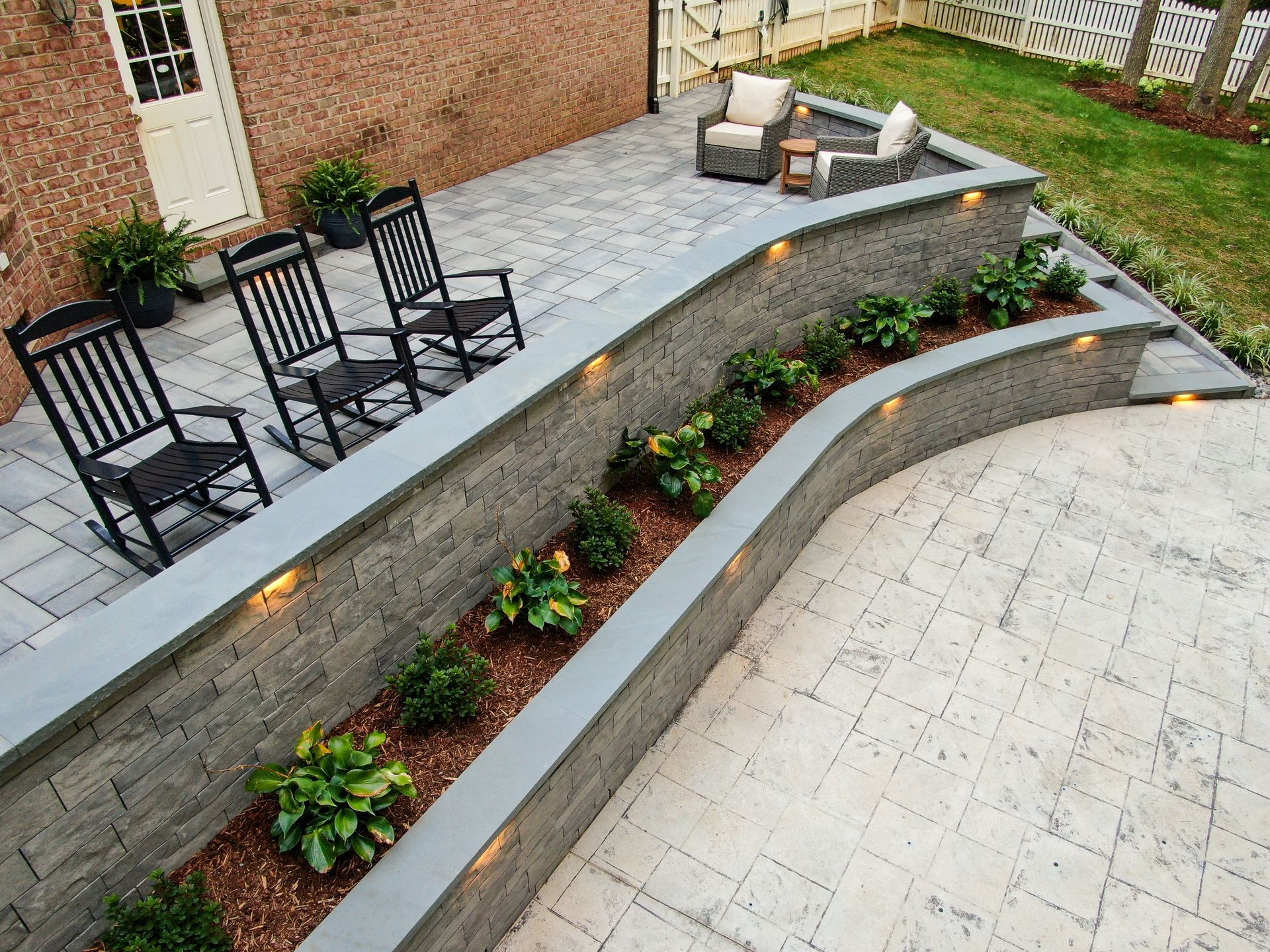 Tiered outdoor patio with black chairs, stone retaining walls, and flower beds beside a paved courtyard