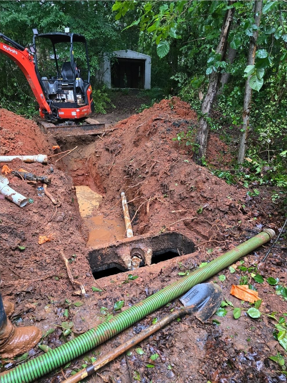 Excavation site with mini-excavator, open septic tank, and green hose. Red dirt, woods in background.
