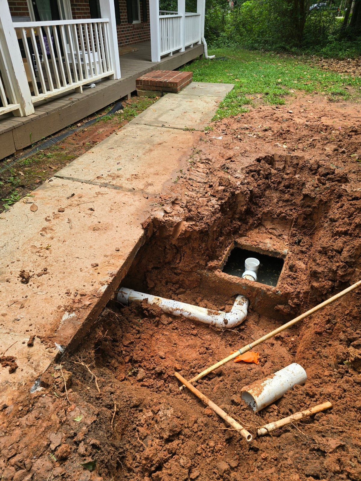 Trench dug in yard exposes white PVC pipes and a square opening near a concrete walkway and house.