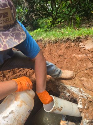 Man in orange gloves connecting a white pipe in a trench; dirt and greenery in the background.