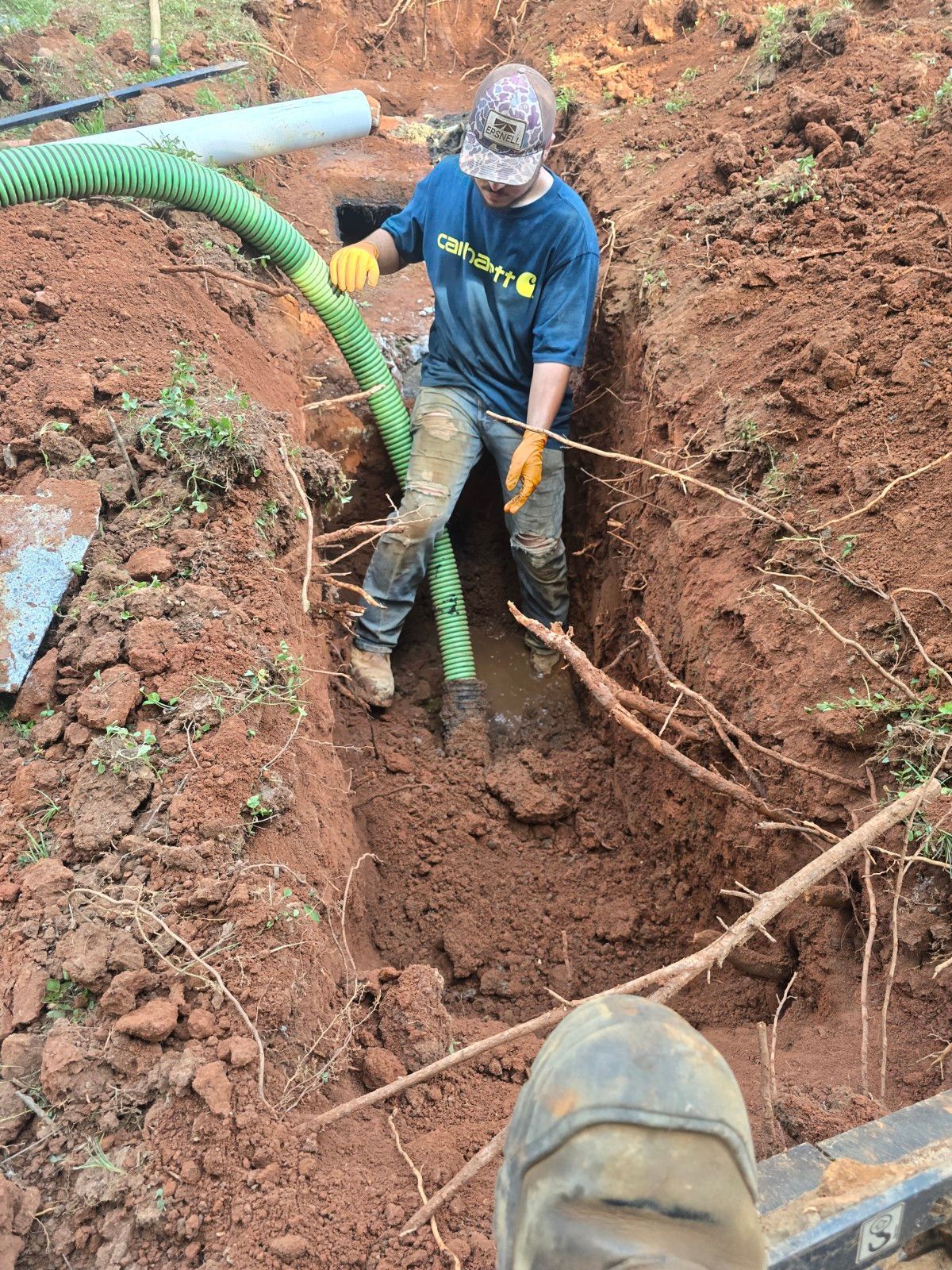 Man in blue shirt works in a trench with a green hose, brown dirt and roots visible.