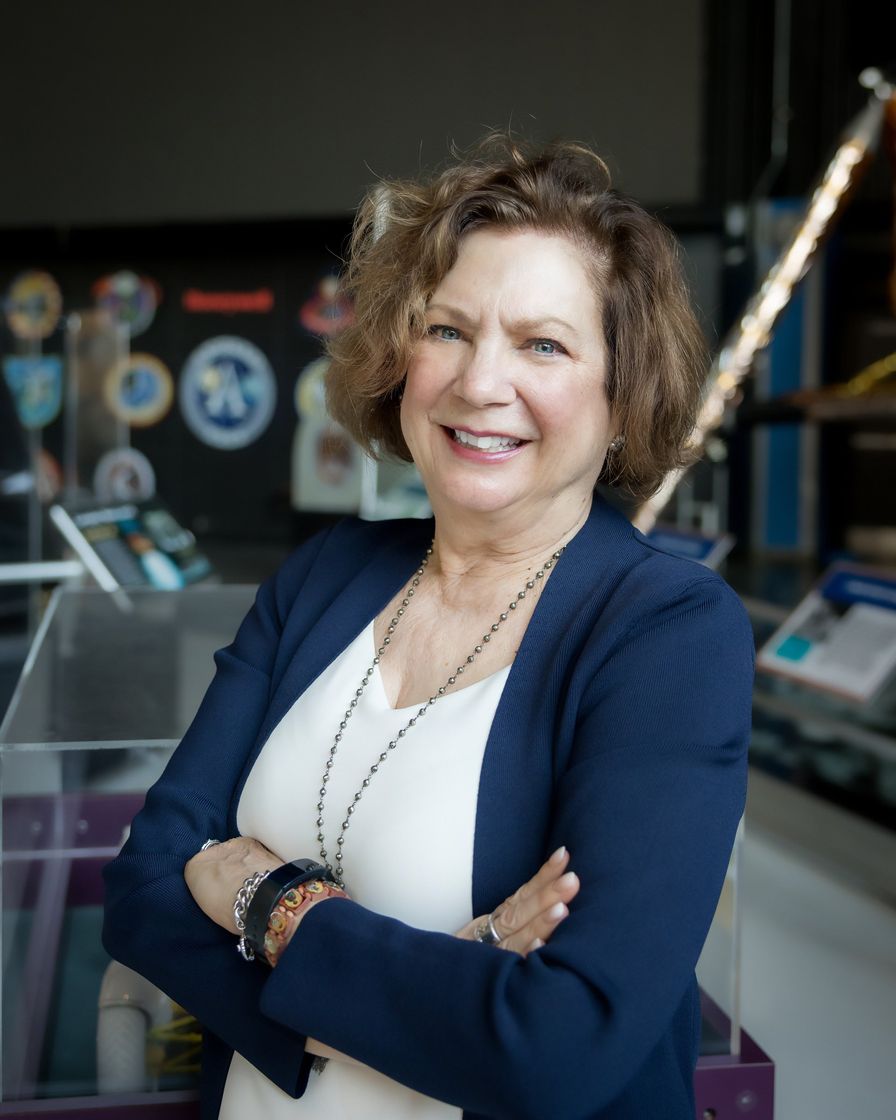 Woman in a navy blazer smiles with arms crossed, in a museum setting, looking at the camera.