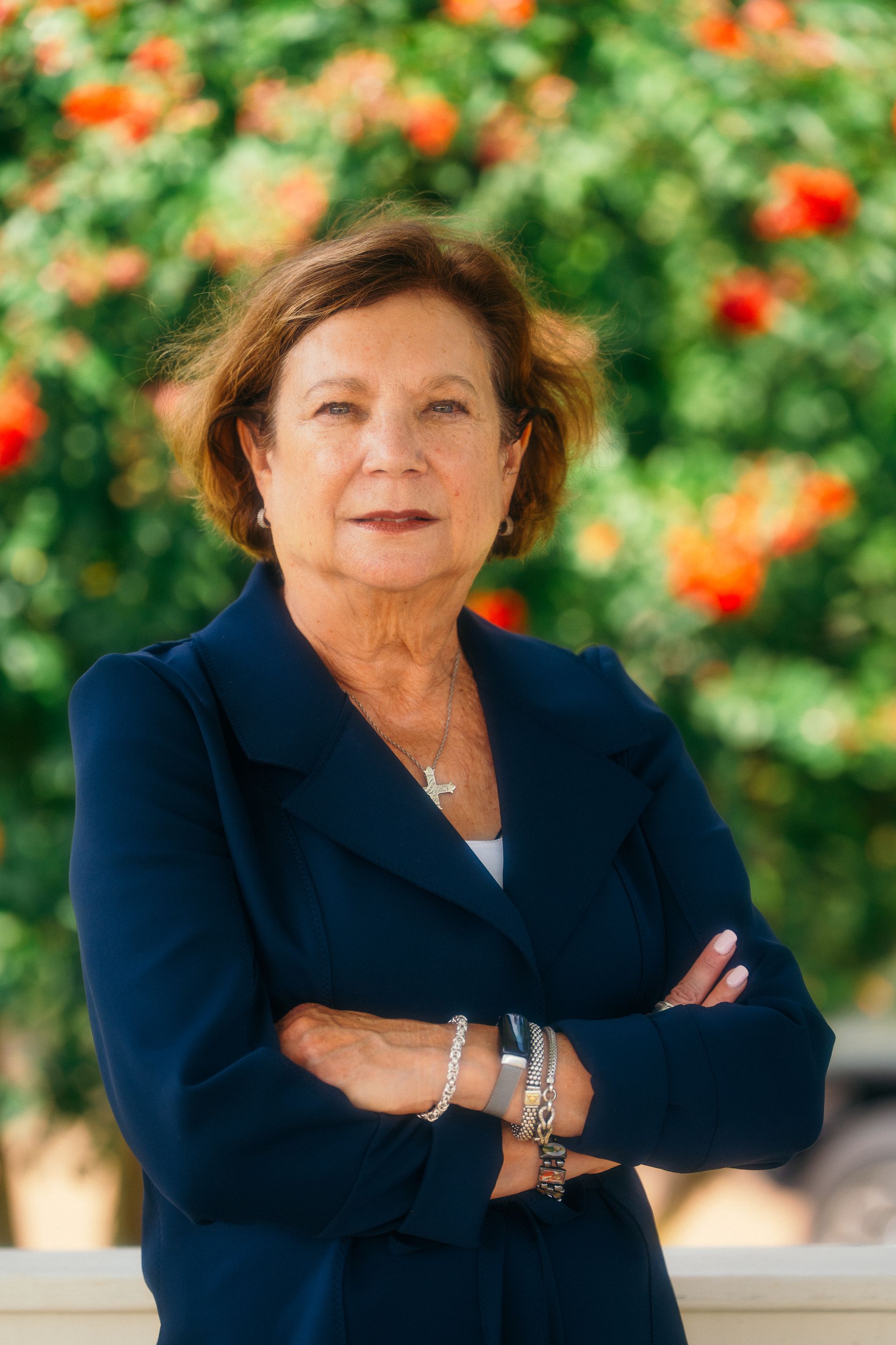 Portrait of a person in a navy blazer standing with arms crossed against a blurred floral background