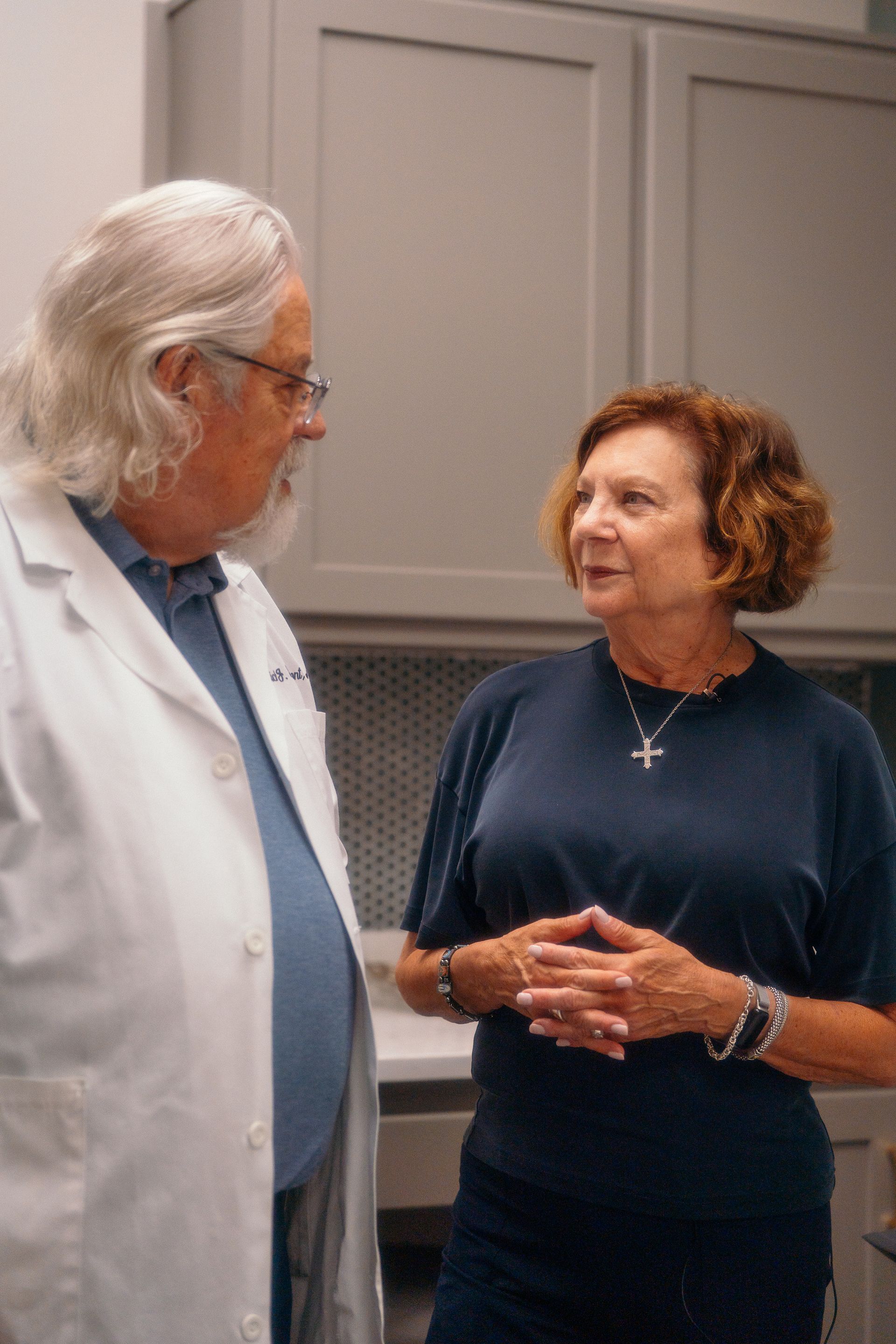 Two people talking in a kitchen, one in a white coat and the other in a dark dress.