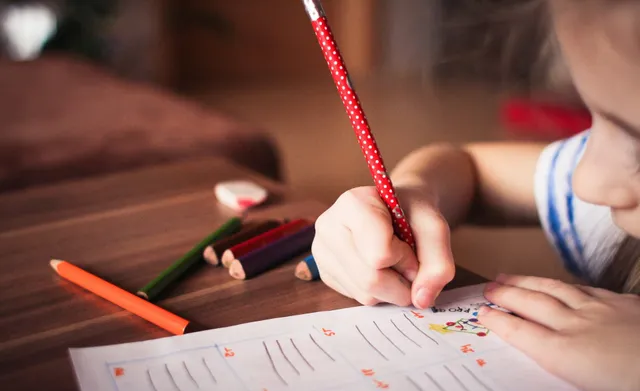 Child's hand holding a red pencil, writing on a worksheet with colorful pencils on a wooden table.