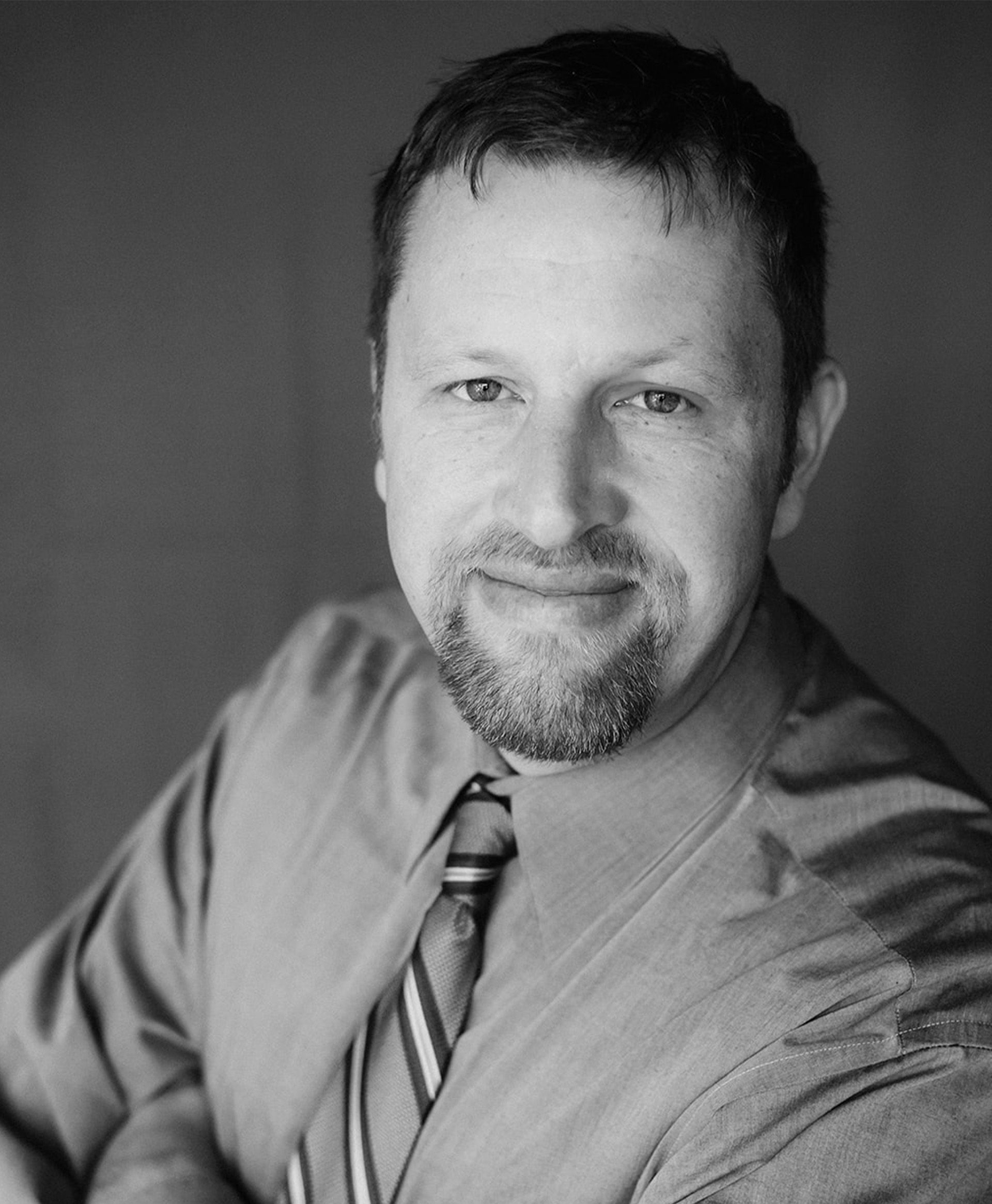 A man in a grey shirt is standing with his arms crossed and smiling.