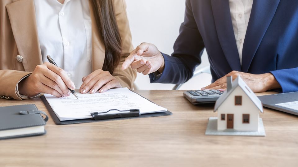 Two women are sitting at a table with a model house and a calculator.