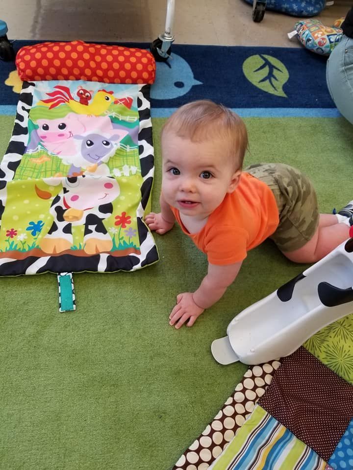 Baby crawling towards the camera on a green rug, next to a cow-themed playmat. The baby wears orange and camo.