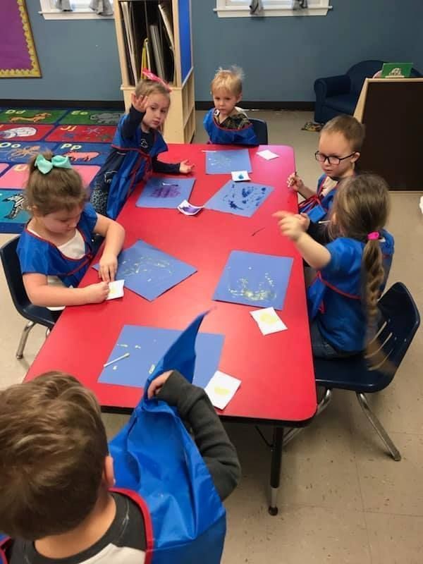 Children in blue shirts at a red table painting on blue paper in a classroom.
