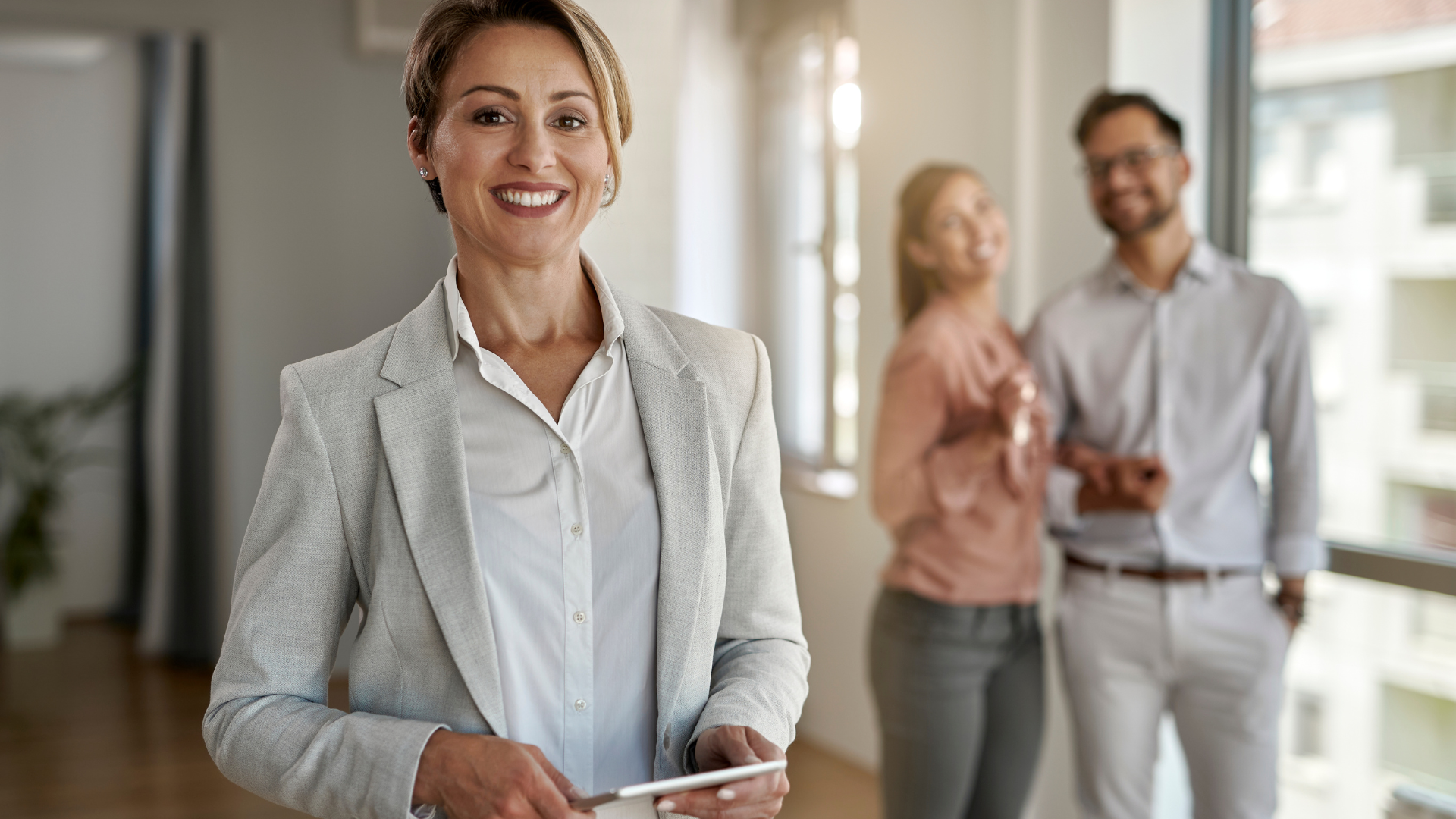 Real estate agent smiles, stands in an empty room, a couple in the background.
