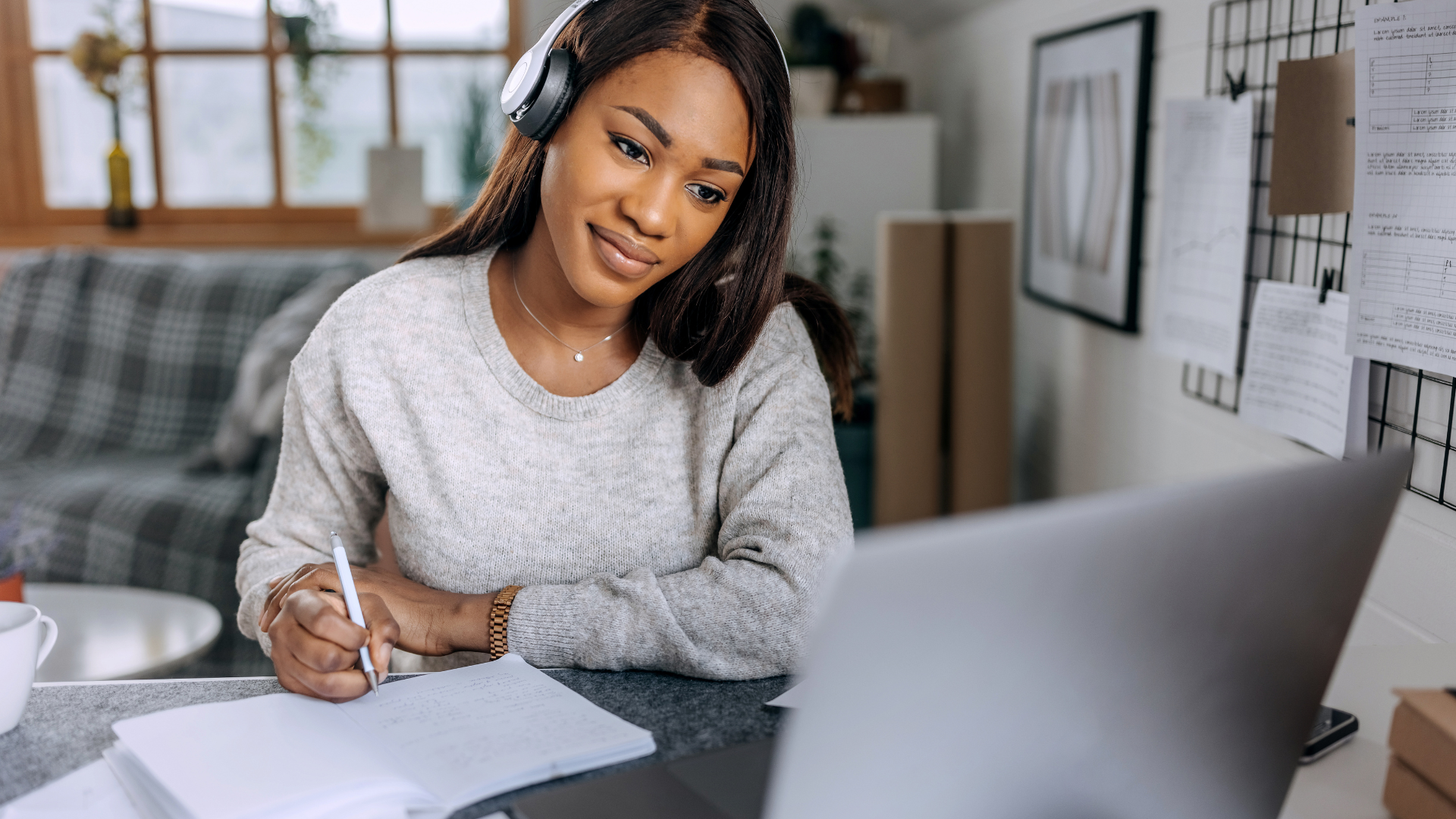 Woman with headphones writing in notebook while looking at laptop.