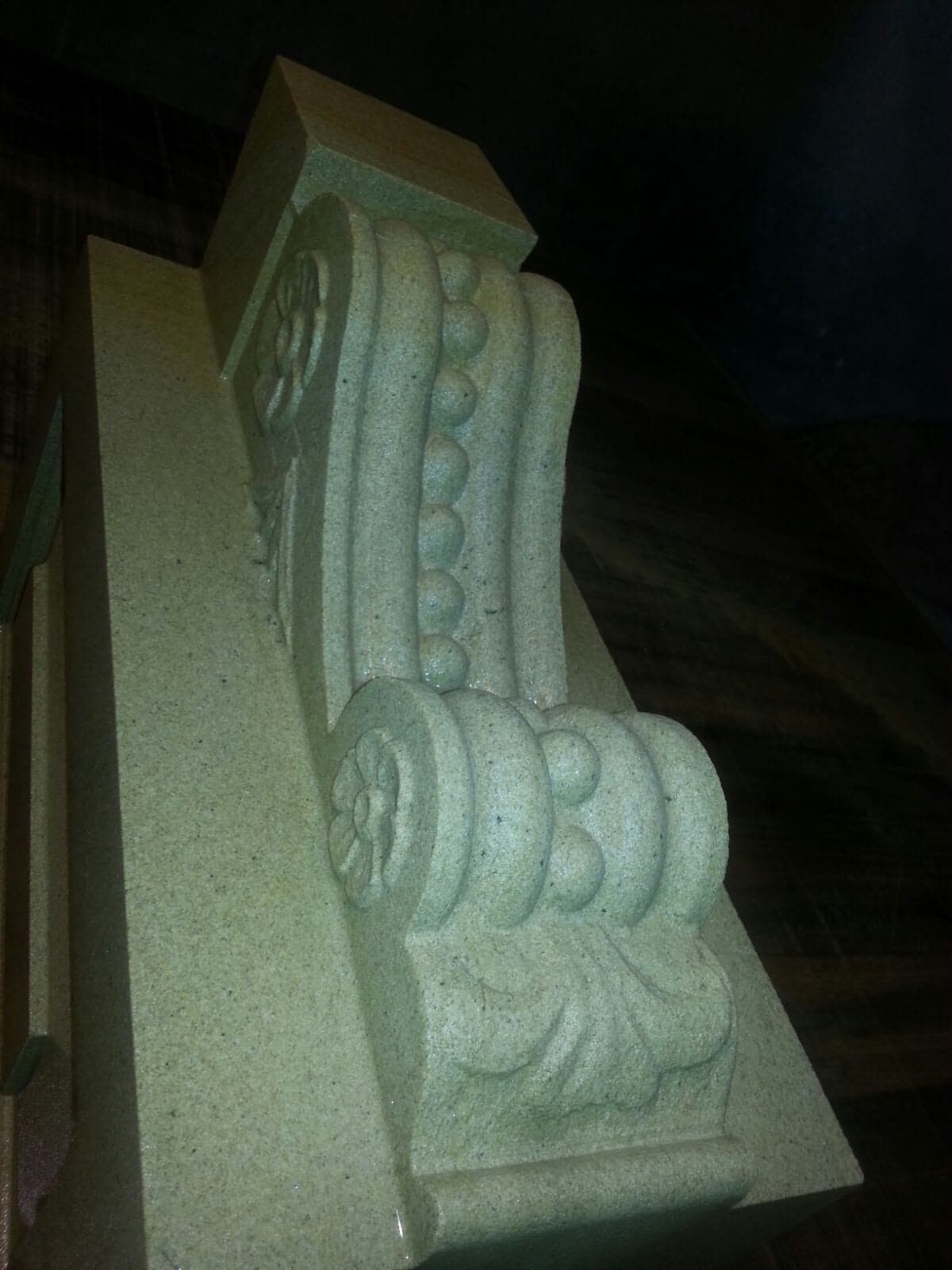 Stone architectural column capital with ornate carved scroll and leaf details, shown close-up against a dark background