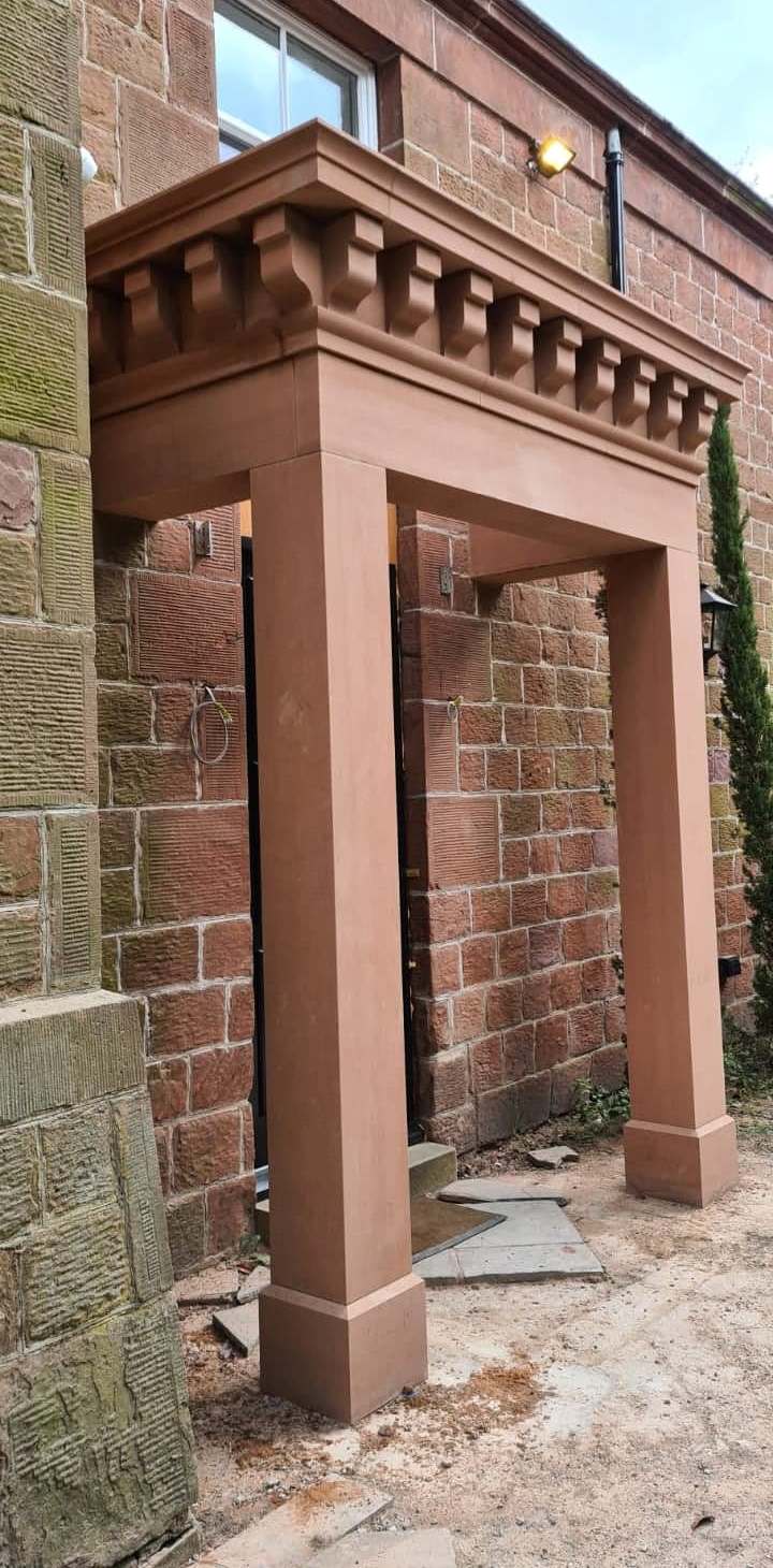 Brown brick entryway structure with two square columns and decorative trim beside a brick building.