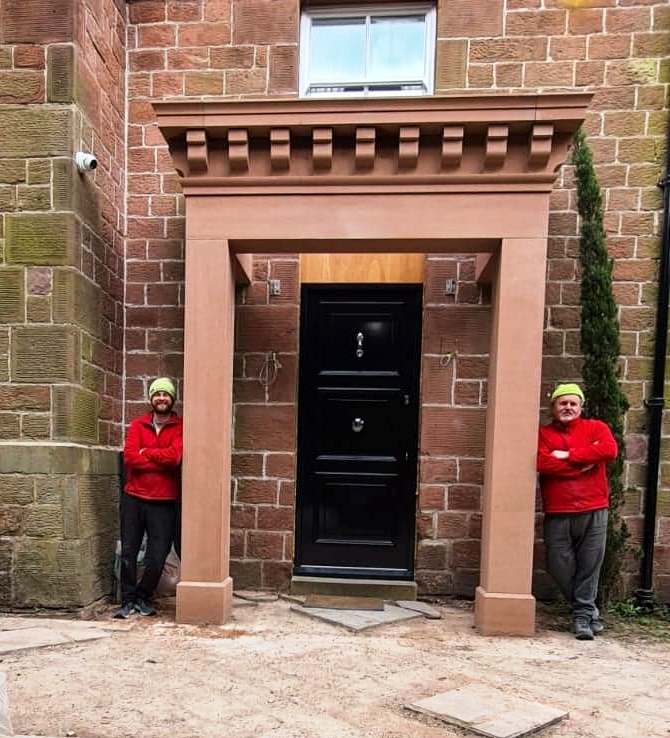 Two workers in red jackets flank a large stone doorway with a black door and brick wall backdrop.