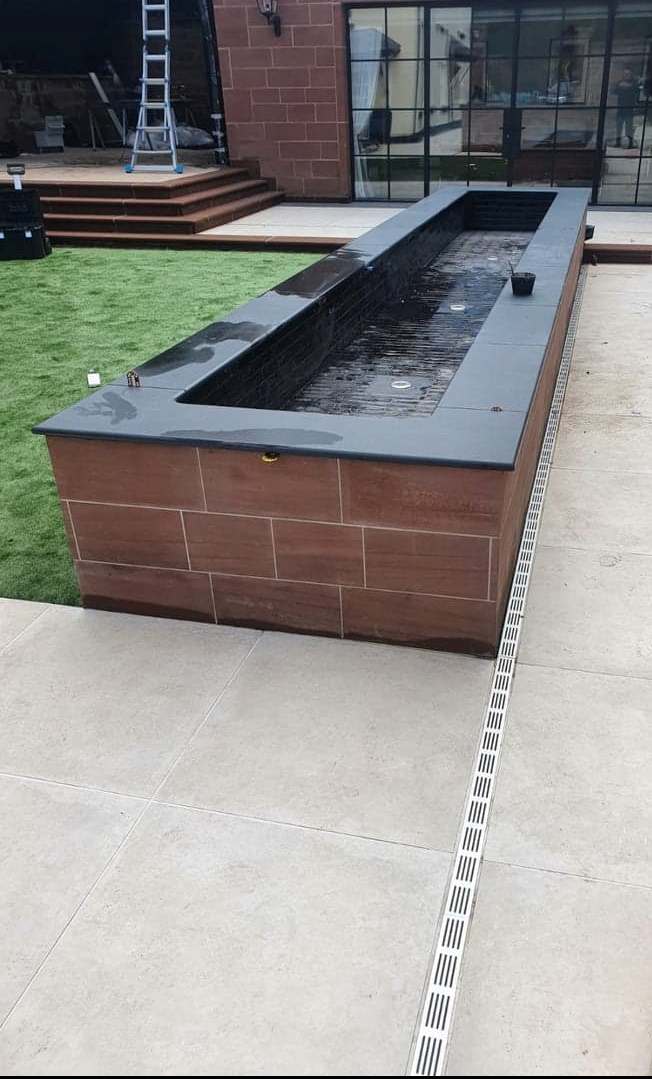 Long rectangular brick planter with black top in a courtyard beside grass and a walkway.