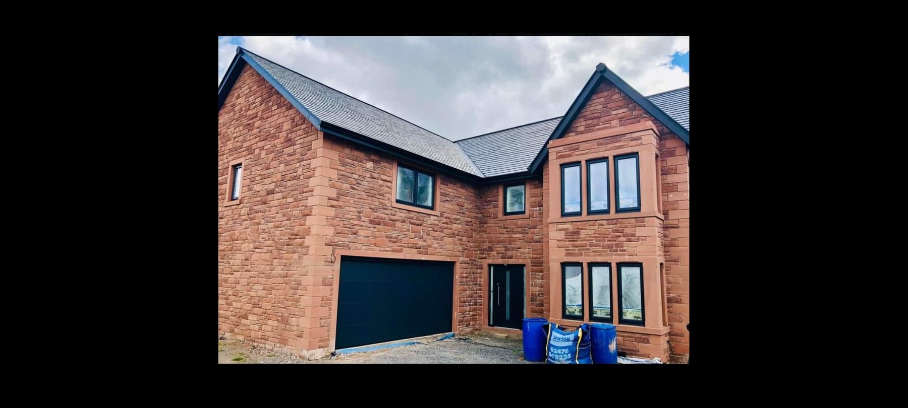 Brick house exterior with black garage door, blue bins, and multiple windows under a cloudy sky