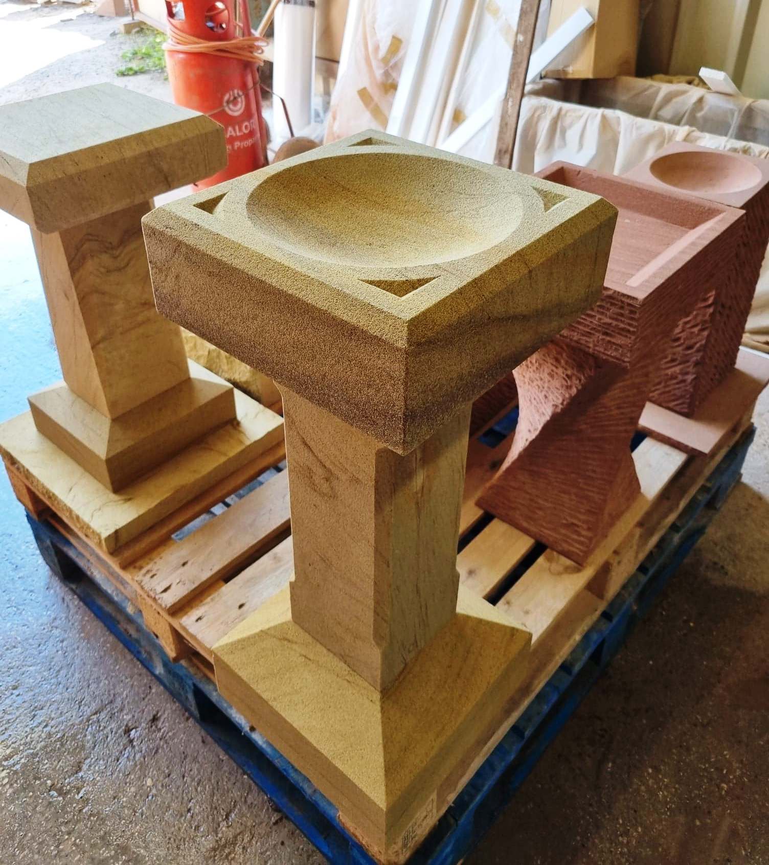 Three stone pedestal sinks on a pallet in a workshop, with tan and pink stone finishes.