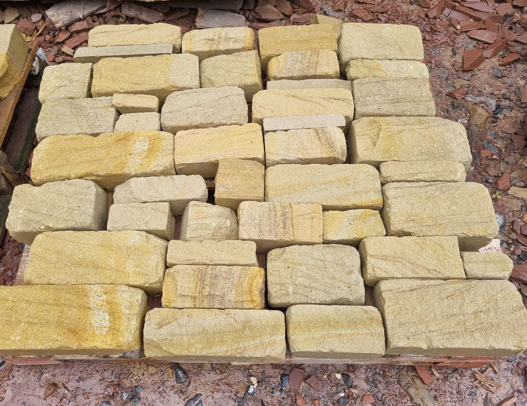 Stacked yellowish stone blocks on the ground outdoors, arranged in rows on dirt and leaves.