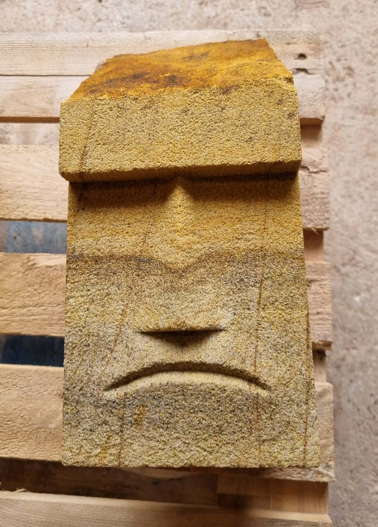 Weathered stone face sculpture with a large brow and a downturned mouth on a wall, yellow-brown in color