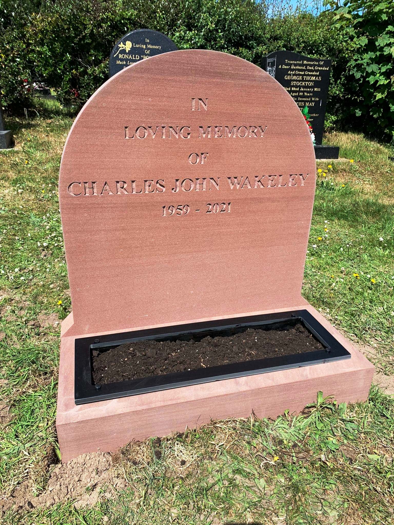 Pink granite memorial stone in grass with engraved text and a black plaque at the base