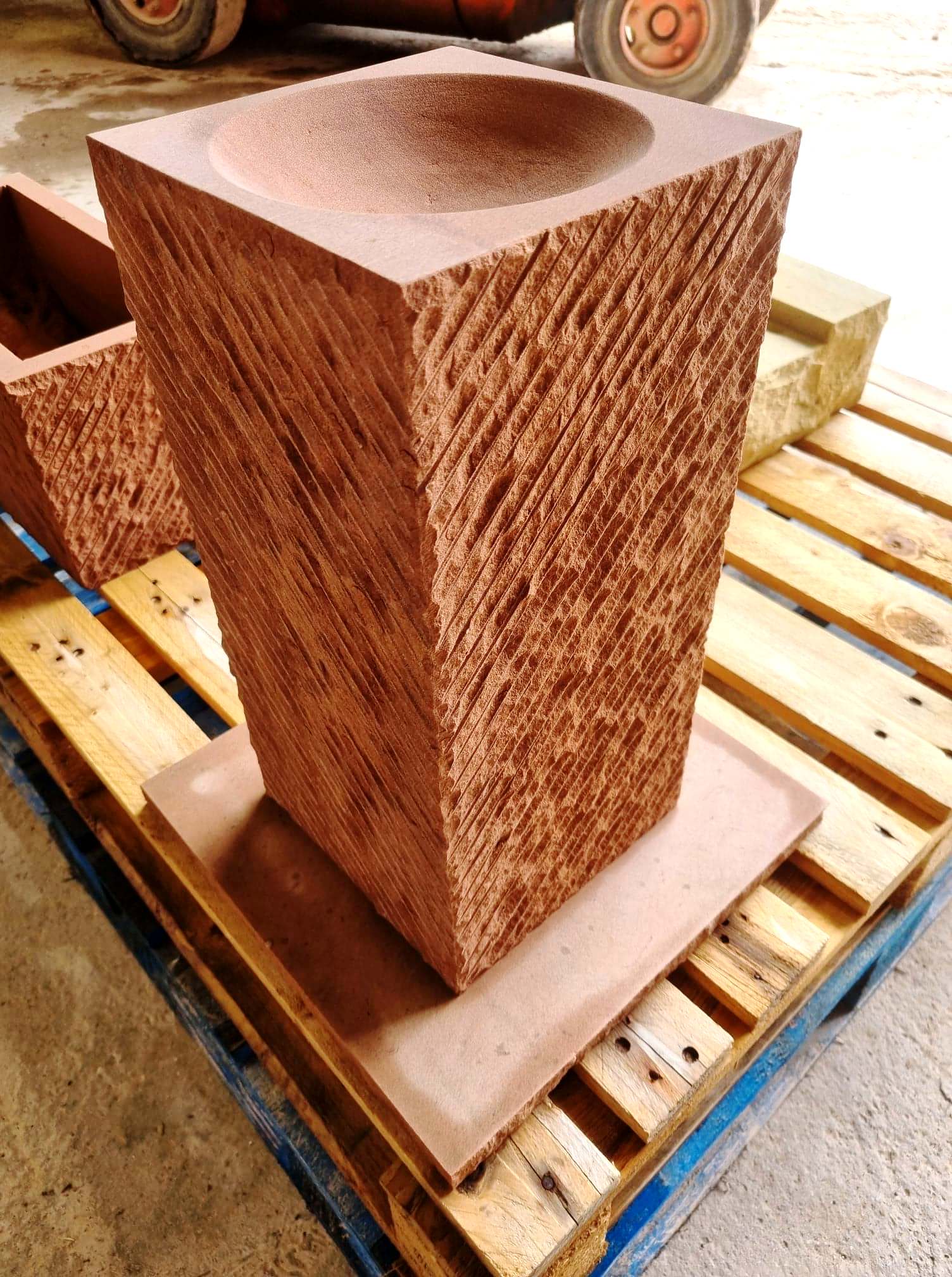 Tall rust-colored stone pedestal on a wooden pallet, with a round recessed top in an outdoor yard.