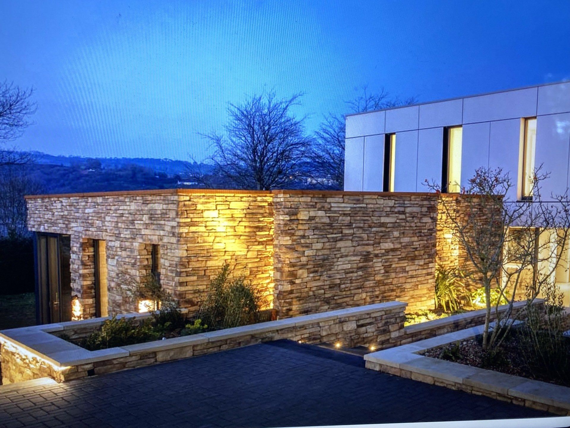 Modern stone house exterior at dusk with warm landscape lighting and blue evening sky