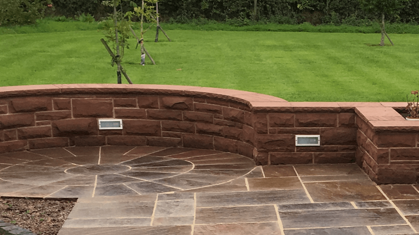 Curved red stone retaining wall with built-in lights beside a paved patio and green lawn.