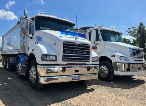 Two mack trucks are parked next to each other on a dirt road.