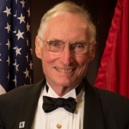 A man in a tuxedo and bow tie stands in front of an american flag