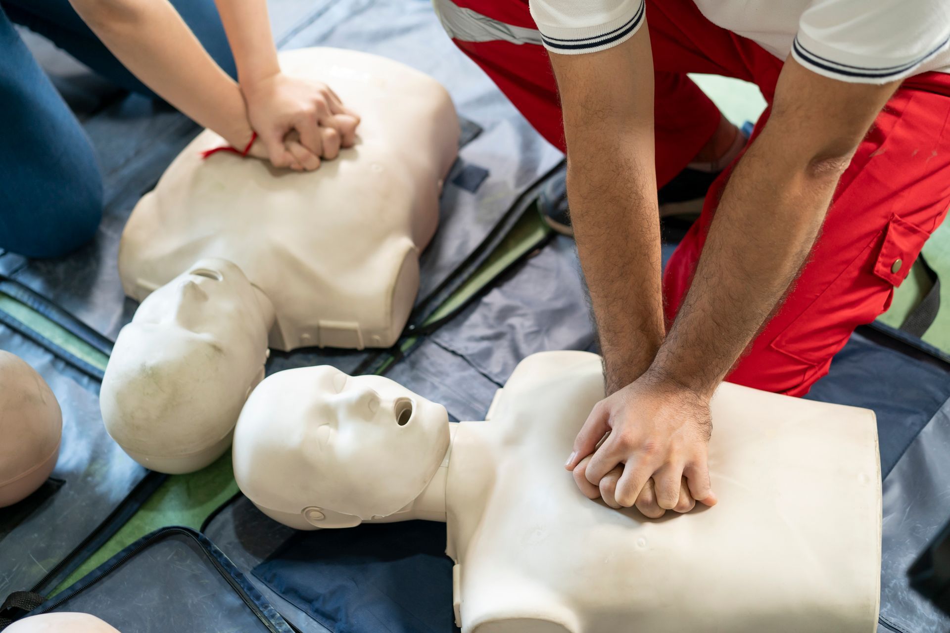 View of a first aid instructor showing CPR to a person on dummies on the ground.