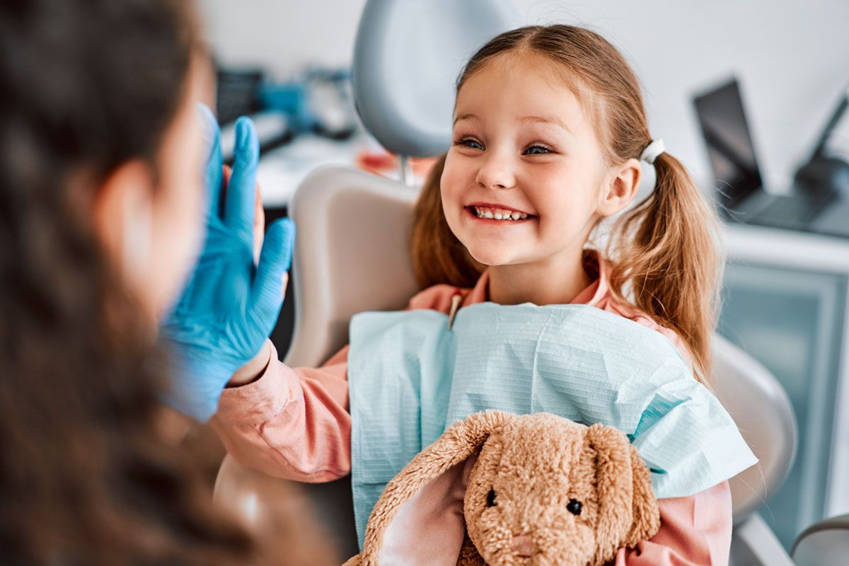 Smiling child in a dentist chair holding a teddy bear, about to high-five a gloved dentist