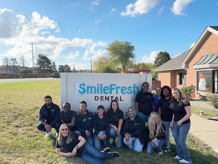 Smile Fresh Dental in Grand Blanc, MI, staff posing outdoors beside dental office sign on a sunny day