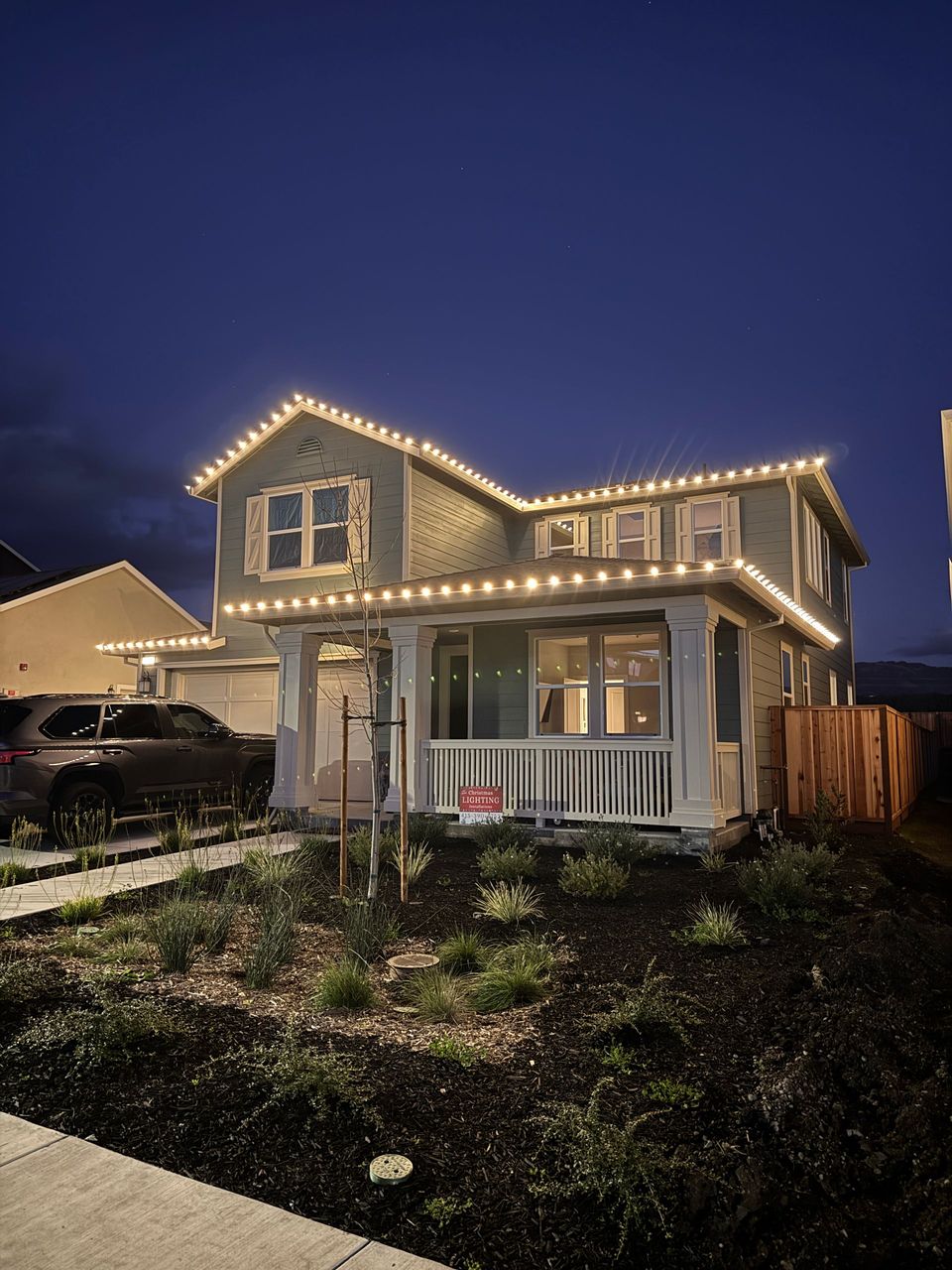 Two-story house at dusk outlined with white string lights in a suburban yard